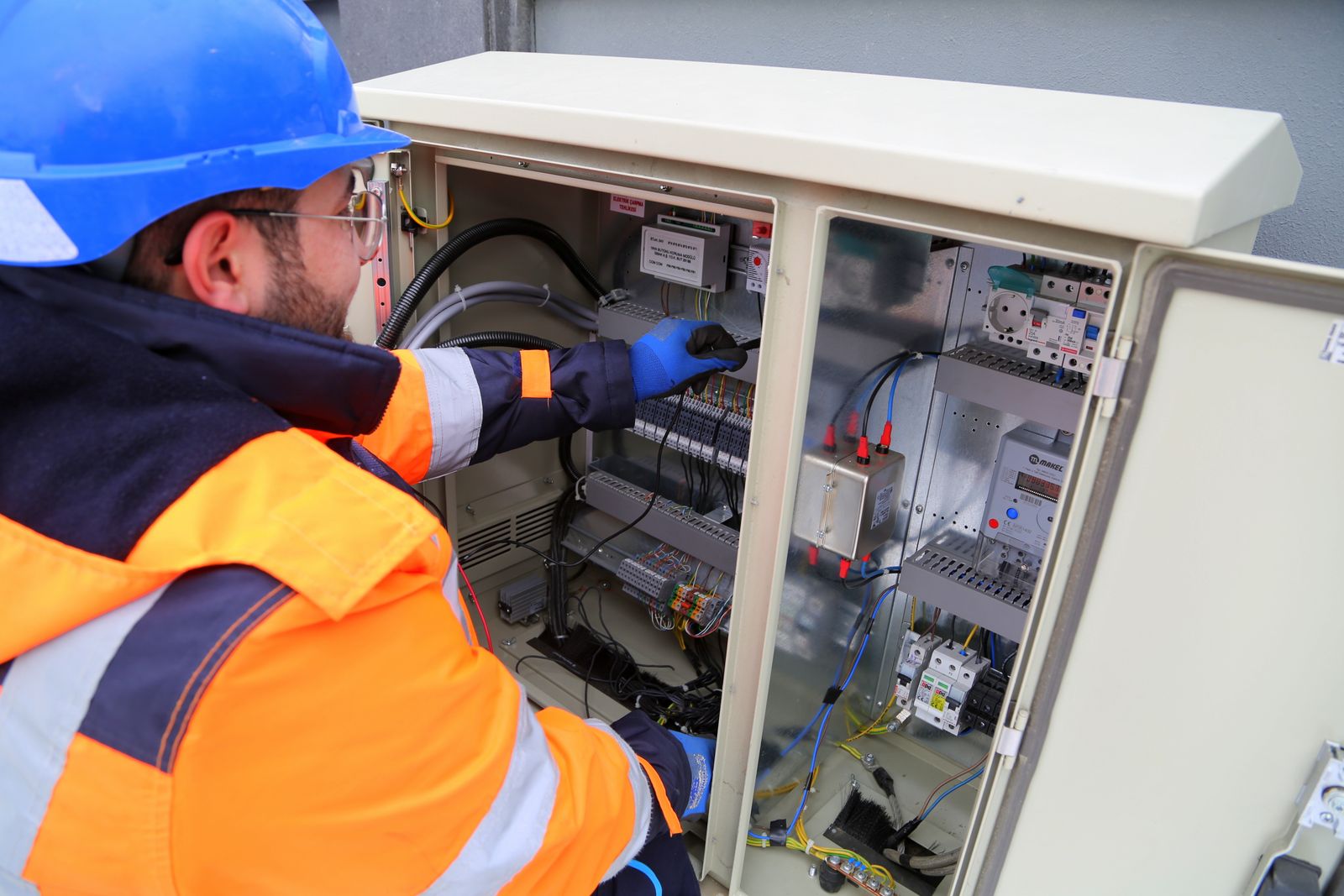 Un instalador con casco y guantes trabaja en un cuadro eléctrico exterior.